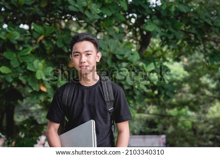 A young college student with a backpack and carrying his laptop while outside the school campus.