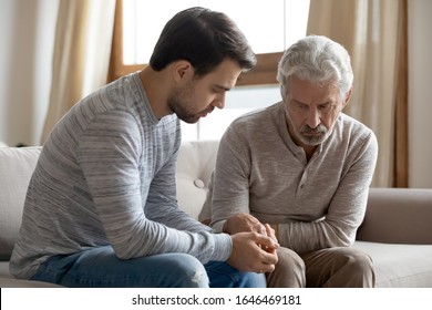 Young Compassionate Man Holding Hand Of Unhappy Elderly Senior Father, Sitting Together On Couch. Worried Grownup Son Supporting Stressed Unhealthy Frustrated Older Dad, Giving Psychological Help.