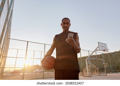 Young concentrated african sportsman holding basketball ball and using mobile phone on sports court. Black man wearing sportswear. Urban basketball player. Sea behind mesh fence. Sunny daytime - Powered by Shutterstock