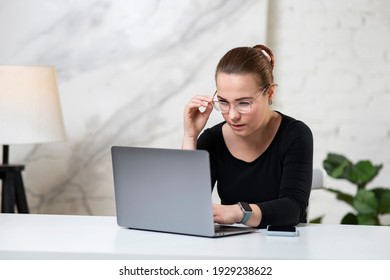 Young Concentrated Focused Girl, Serious Beautiful Woman In Glasses Is Typing, Working On Her Laptop Computer At Table At Home With Eyesight Problems, Hardly Trying To Read Online Content At Screen. 