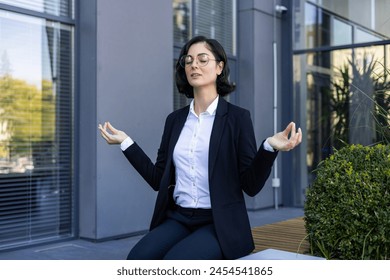 Young concentrated woman in business suit sitting on bench near office desk in yoga pose and meditating, resting and relaxing during work break. - Powered by Shutterstock