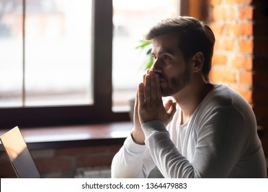 Young concerned businesswoman sitting at table near laptop folding hands together feels doubt unsure thinking making decision or waiting message good news having strong wish hope to get or win concept