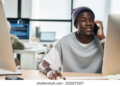 Young Confident African American Software Developer Consulting Client On Mobile Phone While Sitting In Front Of Computer Monitor In Office