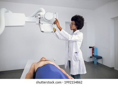 Young Confident Afro American Female Doctor Radiologist, Taking X-ray Scan Of Young Male Patient Lying On The Machine Table. Doctor Standing Near The Patient During Chest X Ray Procedure