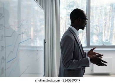 Young Confident Black Man In Grey Suit Explaining Information To Audience While Standing By Whiteboard With Graphs