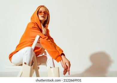 Young confident blonde girl wearing trendy orange hoodie, color sunglasses, posing on white background. Studio fashion portrait. Copy, empty space for text - Shutterstock ID 2122669931
