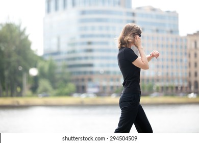 Young confident business woman walking in hurry, looking at watch, talking on mobile phone on the city street in front of blue glass modern office building beside riverbank, profile view
