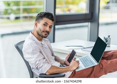 young, confident businessman holding laptop and smartphone with blank screen while looking at camera - Shutterstock ID 1567434298