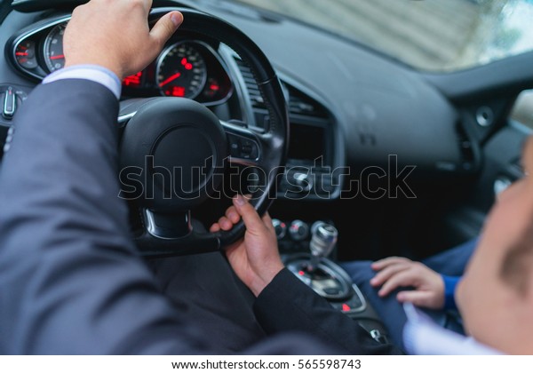 Young confident\
businessman operates a powerful sports car. The bright display and\
steering wheel, sports car interior with a driver in a business\
suit and a passenger\
inside.