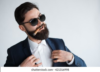 Young Confident Businessman Smoking A Cigar And Fixing His Black Suit With Confidence On White Background. Arrogant Rich Bearded Man In Sunglasses