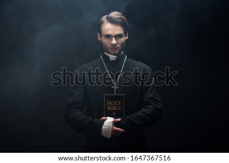 young confident catholic priest looking at camera while holding holy bible on black background with smoke