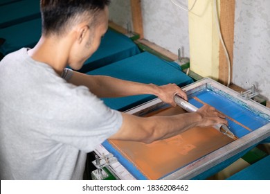 Young Confident, Experienced Male Working With Squeegee On A Small Factory, Handsome Worker Man Using Printmaking Tools, Screen Printing On Clothing Fabric Technique