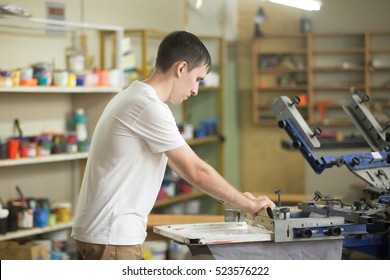 Young Confident, Experienced Male Working With Squeegee On A Small Factory, Handsome Worker Man Using Printmaking Tools, Screen Printing On Clothing Fabric Technique 