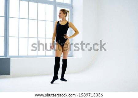 Young confident female choreography with blond hair and pigtail hairstyle wearing black gymnastic costume and knee socks standing in white studio and looking away