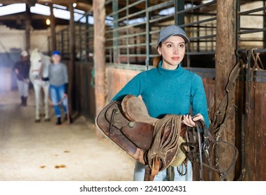 Young confident female stable keeper carrying horse saddle and harness for horseback ride..