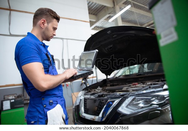 Young\
confident handsome car mechanic with laptop in hand conducts\
computer diagnostics for errors in the car\
systems