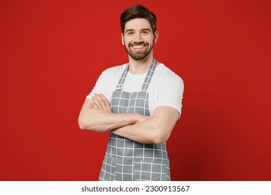 Young confident happy smiling fun male housewife housekeeper chef cook baker man in grey apron hold hands crossed folded look camera isolated on plain red color background studio. Cooking food concept - Powered by Shutterstock