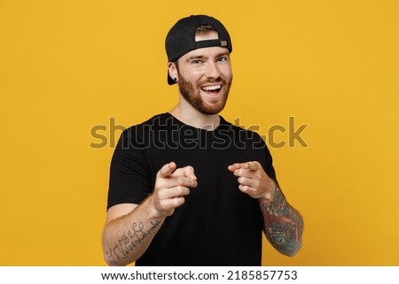 Young confident leader bearded tattooed man 20s he wears casual black t-shirt cap point index finger camera on you motivating encourage isolated on plain yellow wall background. Tattoo translate fun