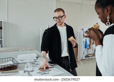 Young confident male fashion designer with snack pointing at one of many sketches of stylish attire while discussing them with colleague - Powered by Shutterstock