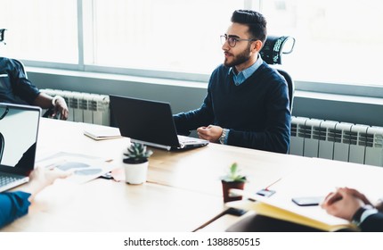 Young confident male manager in optical spectacles sitting at desktop in officce with new modern laptop computer and discussing ideas for IT startup project with colleagues during working time - Shutterstock ID 1388850515