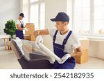 Young confident man mover packing furniture in the living room. Moving service men workers working in clients house wrapping chair with plastic wrap. Move, relocation and moving day concept.