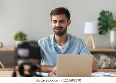 Young confident man speaker talking on digital camera recording vlog sitting at desk with laptop, male vlogger or business trainer speaking shooting blog filming reportage advertising video, coaching