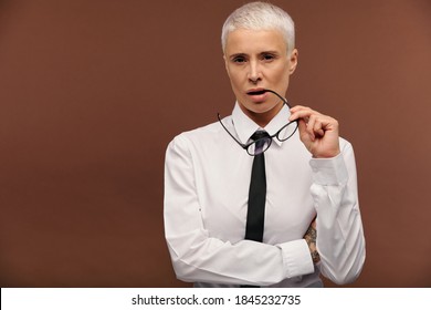 Young Confident Masculine Female In White Shirt And Black Necktie Holding Eyeglasses By Mouth While Standing In Front Of Camera In Isolation