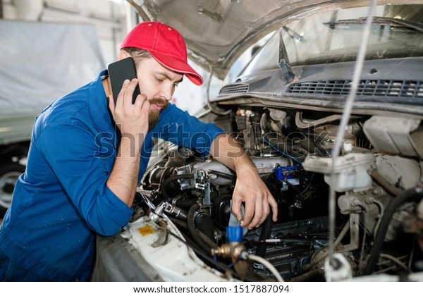 Young confident master of technical service using\
smartphone while consulting one of clients and examining engine of\
machine