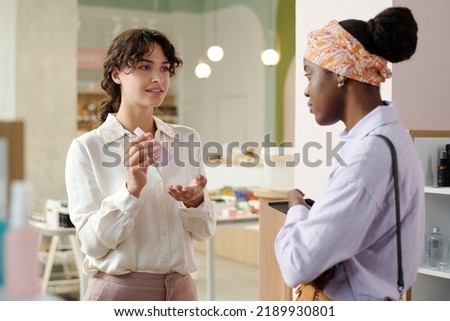 Young confident shop assistant recommending and making presentation of new beauty care product while standing in front of female consumer
