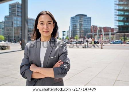 Young confident smiling Asian business woman standing on busy street, portrait. Proud successful female entrepreneur wearing suit posing with arms crossed looking at camera in big city outdoors.