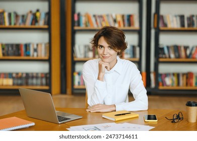 Young confident smiling successful smart employee business woman she wear white shirt casual clothes sit work at library office desk with laptop pc computer, looking camera. Achievement career concept - Powered by Shutterstock