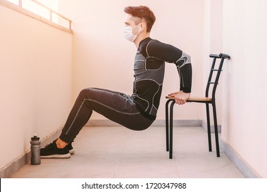 Young confident sports man in medical face mask doing triceps dip on chair at home. Sporty athletic man fitness instructor in protective mask exercising workout for triceps muscle. COVID-19 quarantine