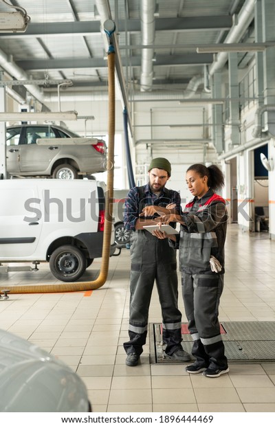 Young confident technicians in workwear\
pointing at one of cars in workshop while discussing its technical\
characteristics before repairing\
it