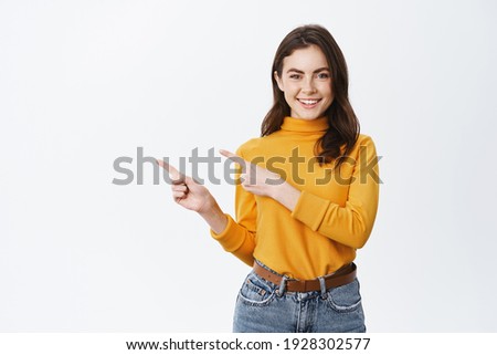 Young confident woman showing logo on empty space. Smiling girl inviting or advertising promo deal, pointing fingers left and looking at camera, white background.