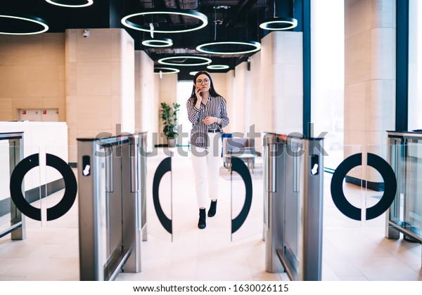 Young confident woman in striped blouse\
and white pants talking on smartphone while leaving modern office\
center through automatic turnstile\
gates