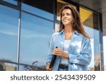 A young, confident woman walking outdoors with a smile, holding a tablet in her hand. She is enjoying a sunny day, standing in front of a modern glass building