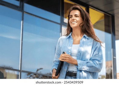 A young, confident woman walking outdoors with a smile, holding a tablet in her hand. She is enjoying a sunny day, standing in front of a modern glass building - Powered by Shutterstock