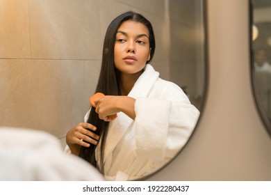 Young confident woman wearing white bathrobe looking at her reflection while standing at the mirror and brush long healthy brunette hair. Millennial female doing morning beauty routine