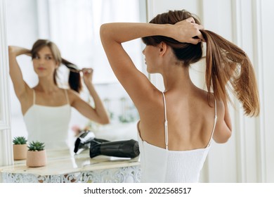 Young confident woman wearing white domestic clothes looking at her reflection while sitting at the mirror and brush long healthy brunette hair. Millennial female doing ponytail at the morning 