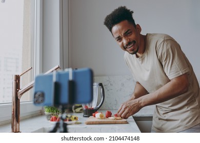 Young Confused African American Man In Casual Clothes Prepare Sweet Breakfast Cut Fruit Watch Series On Mobile Cell Phone Cooking Food In Indoor Kitchen At Home Alone Healthy Diet Lifestyle Concept.