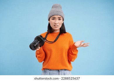 Young confused asian girl in hat, holds digital camera and shrugging shoulders, clueless how to take pictures on digicam, blue background.