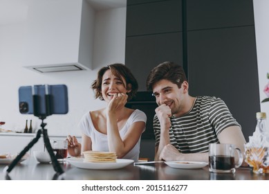 Young Confused Fun Couple Two Woman Man In Casual Clothes Sit By Table Eat Pancake Watch Live Stream By Mobile Cell Phone Prop Up Chin Cook Food In Light Kitchen At Home Together Healthy Diet Concept