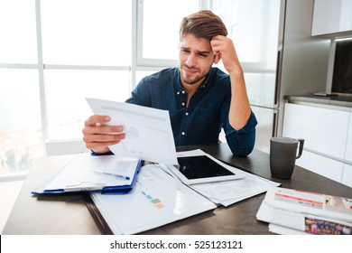 Young confused man analyzing finances at home while holding head with hand and looking at documents. Sitting near table with tablet.