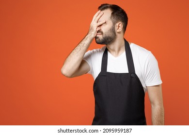 Young confused man barista bartender barman employee in black apron white t-shirt work in coffee shop put hand on face facepalm epic fail gesture isolated on orange background Small business startup