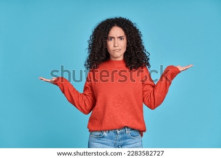 Young confused puzzled latin professional woman, doubtful uncertain hispanic female model student wearing orange sweater standing shrugging thinking of difficult choice isolated on blue background.