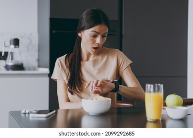 Young confused shcoked sad housewife woman 20s in t-shirt eat breakfast in morning look at smart watch check time hurrying rush cooking food in light kitchen at home Healthy diet lifestyle concept.