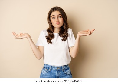 Young Confused Woman Shrugging Shoulders And With Clueless Face Expression, Standing Over Beige Background