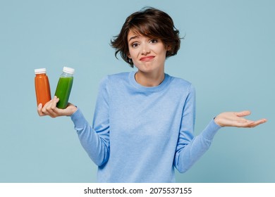 Young confused woman wear casual sweater hold pressed juice green orange vegetable smoothie as detox diet isolated on plain pastel light blue background studio portrait. People lifestyle food concept