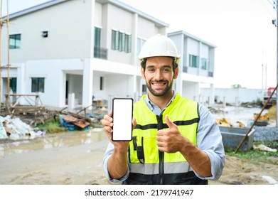 Young Construction Man Smiling Bearded In Vest With White Helmet Demonstrating Blank Smartphone And Showing Thumb Up To The Camera While Standing On Building Construction Site. White Screen, Mockup