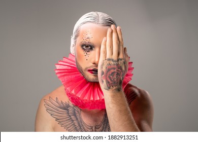 Young Contemporary Actor With Stage Makeup, Tattoo On Chest And Red Paper Jester Collar Around Neck Covering Left Eye By Hand In Isolation